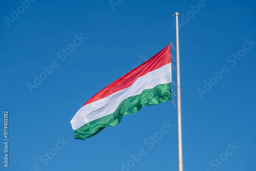 Hungarian national flag waving on a tall flagpole against a clear blue sky, capturing a simple patriotic scene in bright daylight.