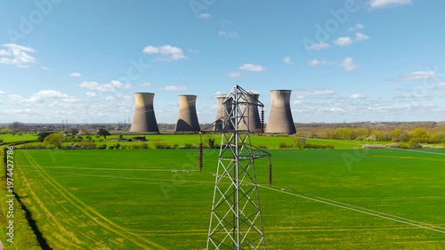 Wide view of power lines crossing green fields toward nuclear power station with multiple cooling towers. Energy infrastructure in rural landscape with transmission network and industrial background