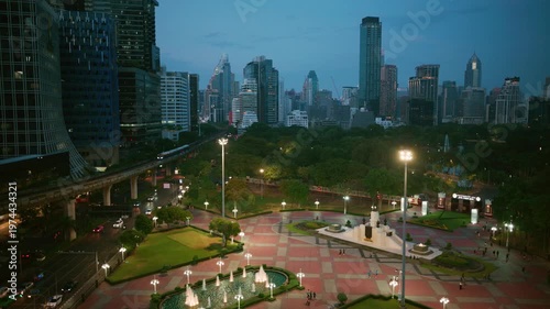 High angle view of Lumphini Park entrance at dusk with illuminated fountains, city skyline, and passing BTS skytrain, Bangkok, Thailand