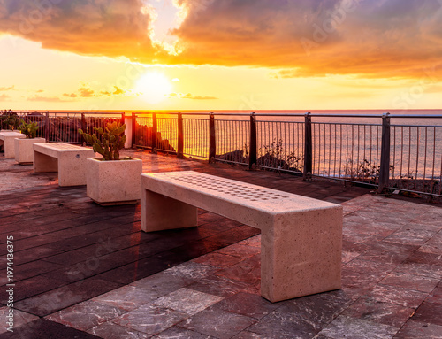beautiful landscape with white stone bench on seafront embarkment during beautiful sunrise or sunset with sidewalk with pavement, sea surf and nice cloudy sunset sky