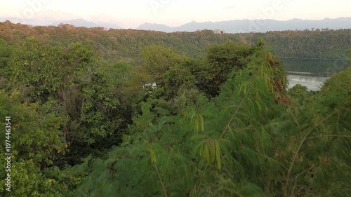 A cinematic drone reveal shot rising over a dirt path and thick vegetation to uncover the hidden Danau Asmara nestled within a dense green valley