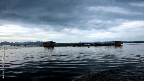 A low-angle point-of-view shot from a moving boat glides across the dark, rippling waters of Cirata Reservoir under a heavy, overcast sky