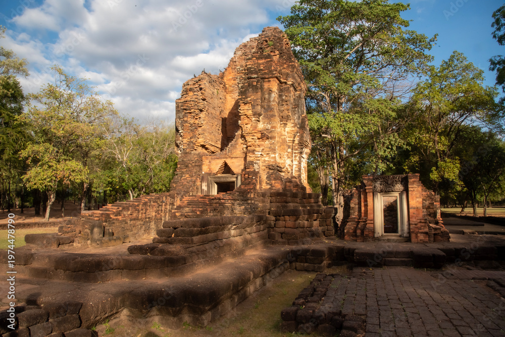 Fototapeta premium Ancient brick ruins at Khao Khlang Nok in Thailand, a historic archaeological site surrounded by trees under a blue sky with clouds