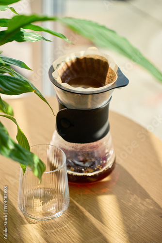 Fresh brewed coffee in filter dripper with empty glass on wooden table in sunlight. Concept of manual brewing home coffee routine and calm morning lifestyle.