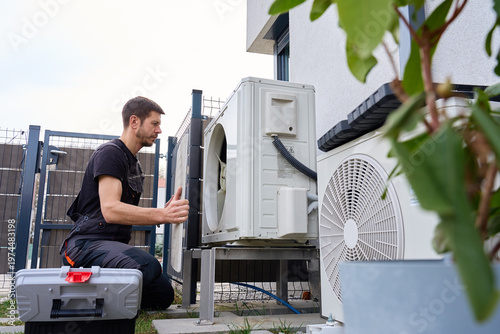 Technician performing maintenance on outdoor heat pump unit near residential building exterior. Man installing air source heat pump using tools. Concept of HVAC service and heating system repair