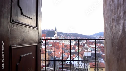 View through open antique wooden doors to a historic old town with red roofs, a medieval church spire, and scenic hills. Cinematic travel and architectural heritage concept.