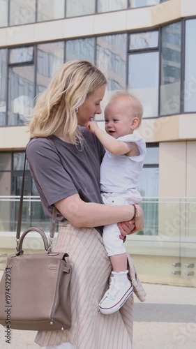 Beautiful mother holding child outdoors in city at summer. Tender love and affection as mom hugs and kisses baby, bright beige tones, warm emotions, modern urban family lifestyle