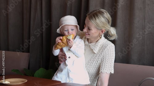 Stylish blonde mom with baby girl in cozy cafe. Happy woman smiles with love while child eats croissant. Motherhood, care and tenderness indoors in modern cafe