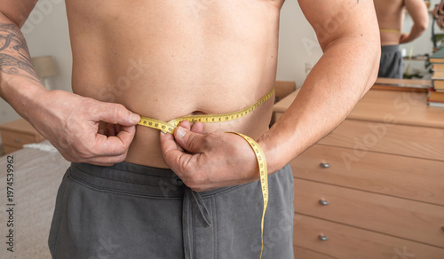 Man measuring his waist with a tape measure, checking progress on his weight loss journey in the bedroom. Real person, self-acceptance, authentic lifestyle