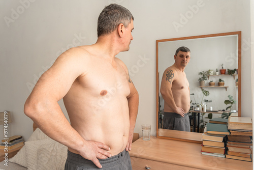Fotografie Mature man standing shirtless, looking at his reflection in a mirror, examining his changing body shape and waistline