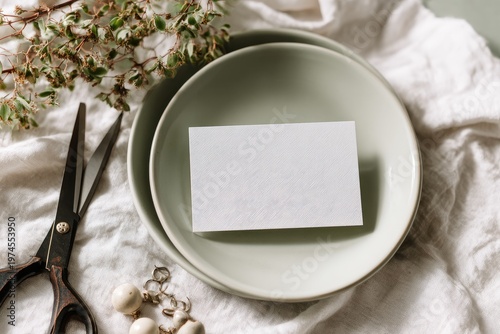 Blank white card on green plate with scissors, sprigs, and linen fabric backdrop