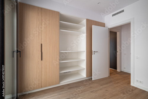 Modern empty room with wooden flooring and a built-in wardrobe. The wardrobe features open shelves and a closed door. Natural light enters through a nearby window.