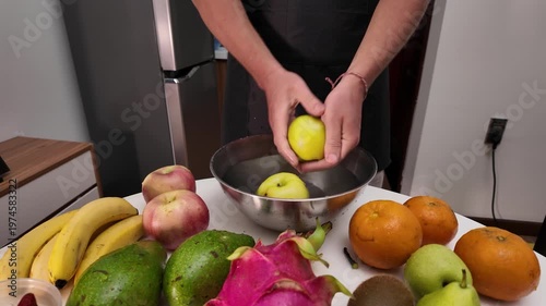 A man in an apron stands in front of a table with fruit, washing green apples in a metal bowl filled with water before cooking. Close-up, front view.