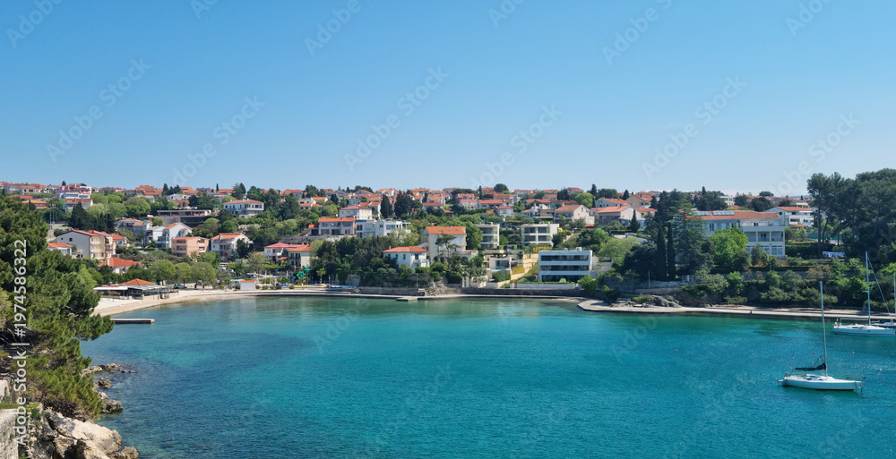 Fototapeta premium A wide scenic view showing the blue sea bay of Krk town with houses and lush green pine trees under a clear sky.