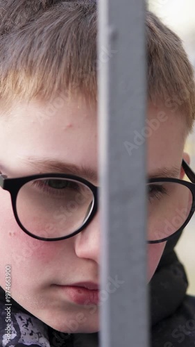 A teenage boy stands behind a metal fence, looking upset and isolated. The scene conveys sadness and bullying at school, highlighting emotional struggle and vulnerability. Vertical video.