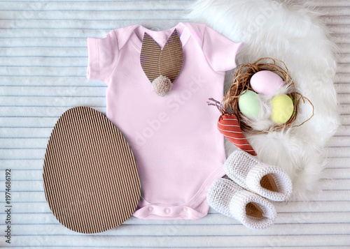 Baby bodysuit with bunny ear decoration next to easter egg, nest with eggs, and knitted booties. Creative flat lay composition for easter holiday baby announcement or celebration.