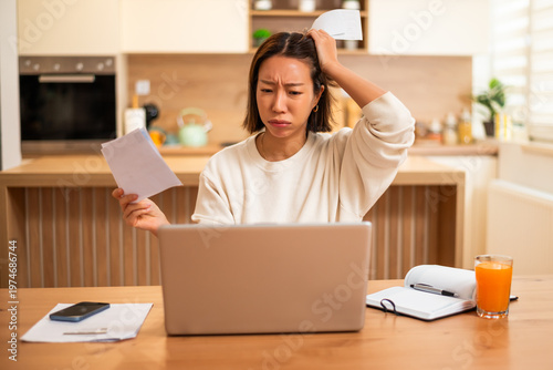 A worried woman sits at a table reviewing bills and documents while looking at her laptop, appearing stressed about her finances.