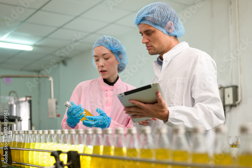 Food quality inspectors checking bottled fruit juice on production line in beverage factory, using tablet for monitoring and quality assurance.