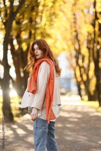 Red-haired woman in autumn park wearing coat, jeans and scarf