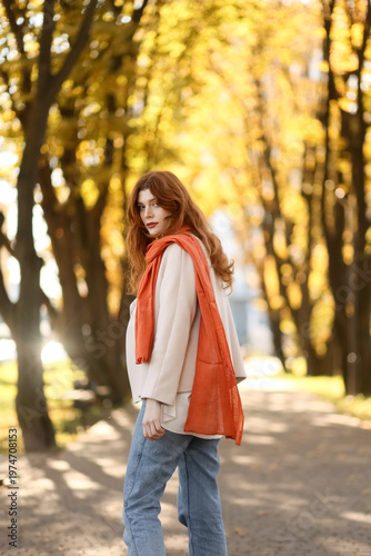 Red-haired woman in autumn park wearing coat, jeans and scarf