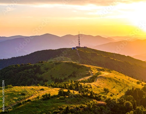 A scenic vista showcasing a vibrant sunset over a mountain range with a communications tower and winding pathways. The landscape is lush