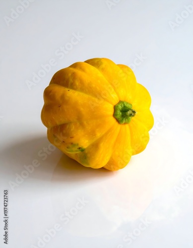 A single, vibrant yellow pattypan squash sits on a white, reflective surface. The squash features a ridged exterior and a green stem