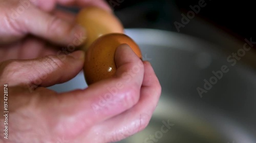 Close up of hands with polished nails gently peeling brown shell off boiled egg over glass bowl, revealing smooth white surface, with soft blur denim and indoor background