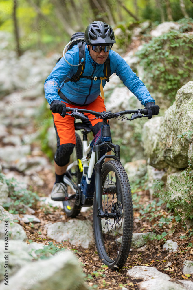 Fototapeta premium Experienced mountain biker riding a rocky forest trail wearing helmet, backpack and bright gear