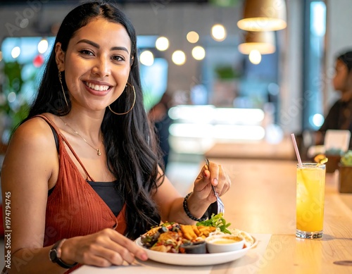 A smiling woman with dark hair enjoys a meal at a restaurant, holding a fork over a plate of food, with a beverage