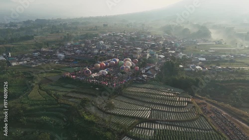 Aerial view of colorful hot air balloons being prepared for launch in a lively field near Wonosobo, with the majestic Mount Sindoro rising in the background.
