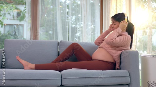 Happy pregnant woman wearing headphone holding smartphone to relax on couch at home enjoying peaceful lifestyle eating healthy yellow banana listening music in cozy living room with bright sunlight