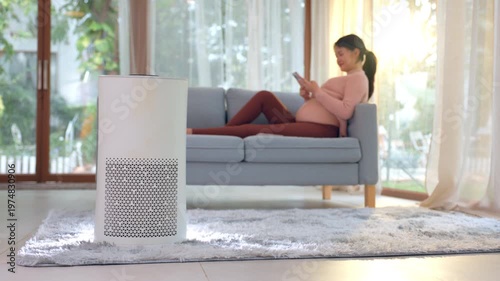 Happy pregnant Asian woman relaxing peacefully on comfortable blue sofa using smartphone near modern white air purifier filtering clean indoor environment enjoying fresh sunlight