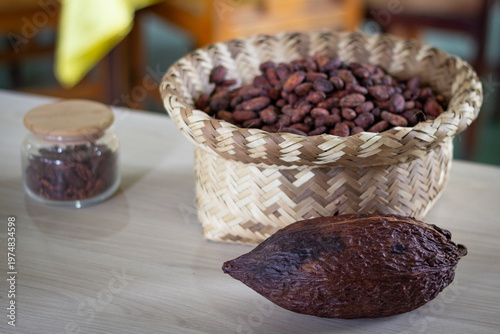 Cacao pod and roasted cacao beans in a woven basket, with raw cacao nibs in a glass jar, displaying the stages of chocolate production on a wooden table