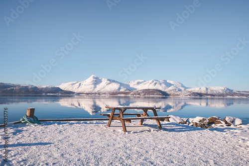 Wooden picnic table in snow overlooking Malangen fjord near Tromso, Norway. Calm water reflects snowy mountains under clear blue sky, creating peaceful Arctic landscape