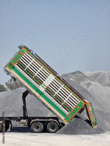 A heavy-duty dump truck with a raised, colorful bed unloading a massive pile of grey gravel at a depot.