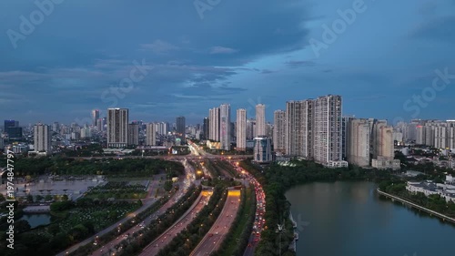 Drone perspective of curving Thang Long Boulevard during blue hour with heavy traffic flow, modern skyline, and lakeside residential buildings in Hanoi
