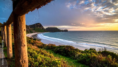 Scenic beach view at sunset, with mountains backdrop. Warm light illuminates waves and greenery near rustic wooden railing