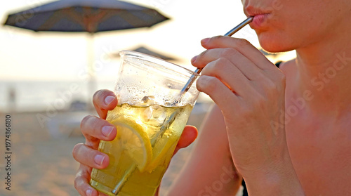Woman drinking iced lemonade with straw on beach during summer. Refreshing citrus beverage in plastic cup. Vacation lifestyle and holiday relaxation at seaside during sunny weather.