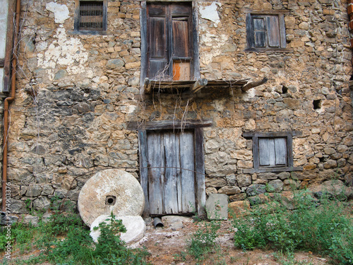 Obraz na płótnie Weathered Stone Mill House with Grinding Stones - Facade of a rustic, abandoned stone building with old wooden doors and two antique millstones outside