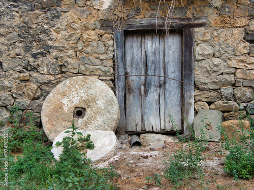 Fotografie Ancient Stone House with Millstones and Doors - Abandoned rural house facade with rustic stone wall, weathered wooden doors and windows, and historic millstones in the foreground