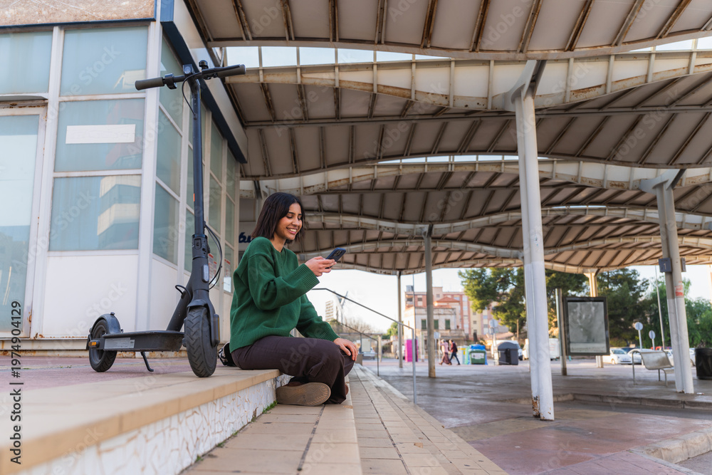 Fototapeta premium Young woman smiling, scrolling smartphone, sitting on steps with her electric scooter in an urban setting