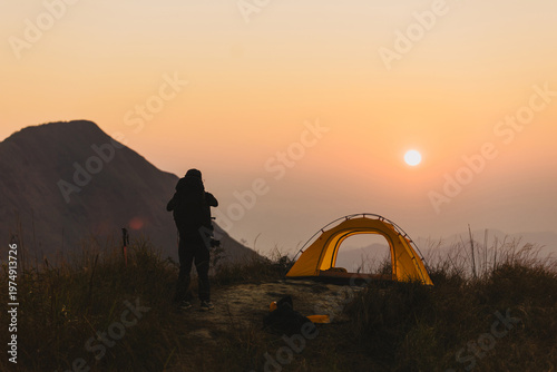 traveler camping with yellow tent on mountain ridge at sunset. Silhouette of man looking at view against majestic peak. Adventure, outdoor lifestyle and trekking concept