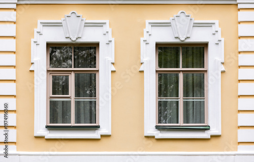 Symmetrical yellow building facade featuring two classic double windows with ornate white trim and stucco walls, subtle reflections in glass, evoking historic European architecture. Background texture