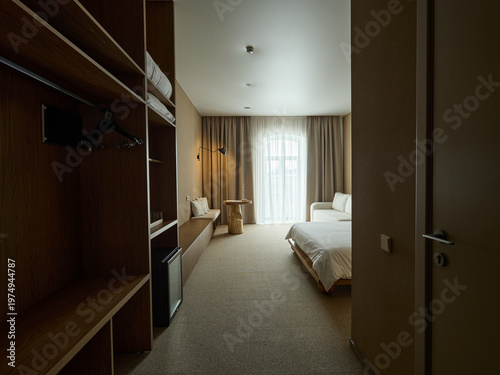 Modern hotel room interior with wooden wardrobe, long cabinet, and twin beds near a large window with sheer curtains. Warm beige tones and minimalist design.