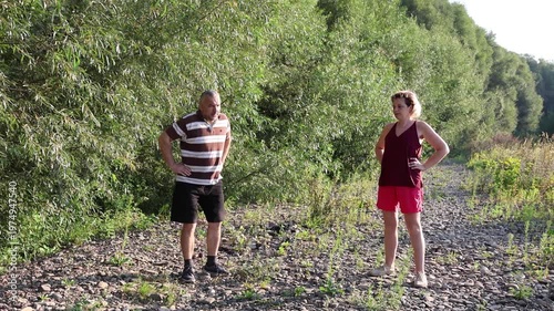 Two people perform morning physical exercises on a rocky riverside surrounded by greenery on a sunny summer day.