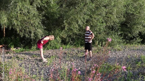 Two people perform morning physical exercises on a rocky riverside surrounded by greenery on a sunny summer day.