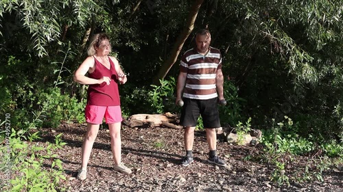 Two people perform morning physical exercises on a rocky riverside surrounded by greenery on a sunny summer day.