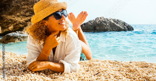 Woman relaxing on a pebble beach wearing sunhat and sunglasses by turquoise sea on summer vacation