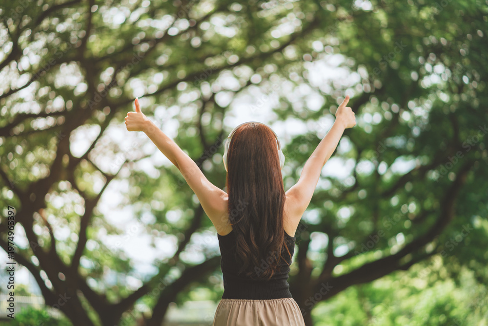 Fototapeta premium Back view of a young woman with headphones raising her thumbs up in a lush green park, celebrating success and freedom through music and nature during a sunny day to promote mental wellness