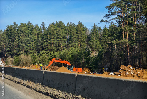 Orange excavator moving earth to form highway embankment while surveyor with total station measures site beside pine forest under clear blue sky during daytime construction.